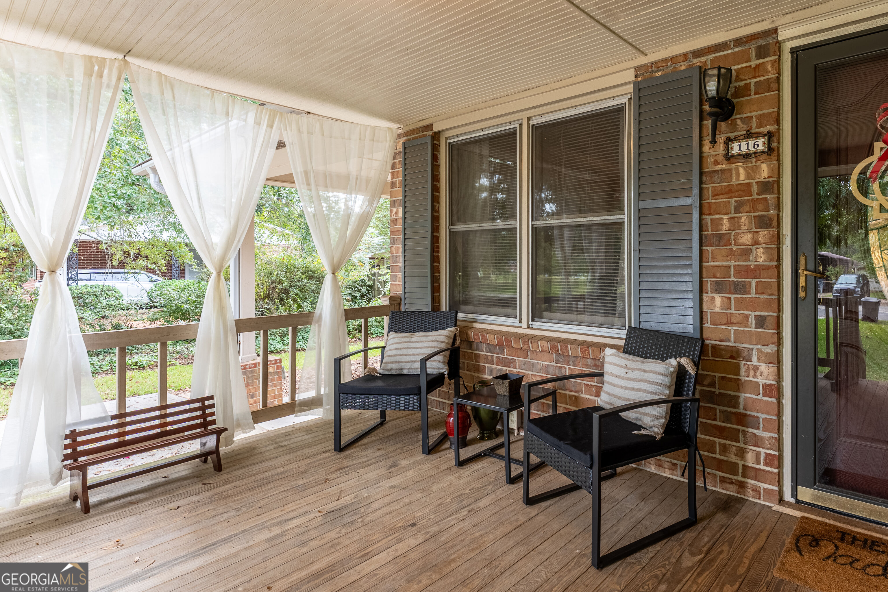 Covered brick front porch with sheer curtains, a wooden bench, and two cushioned armchairs.