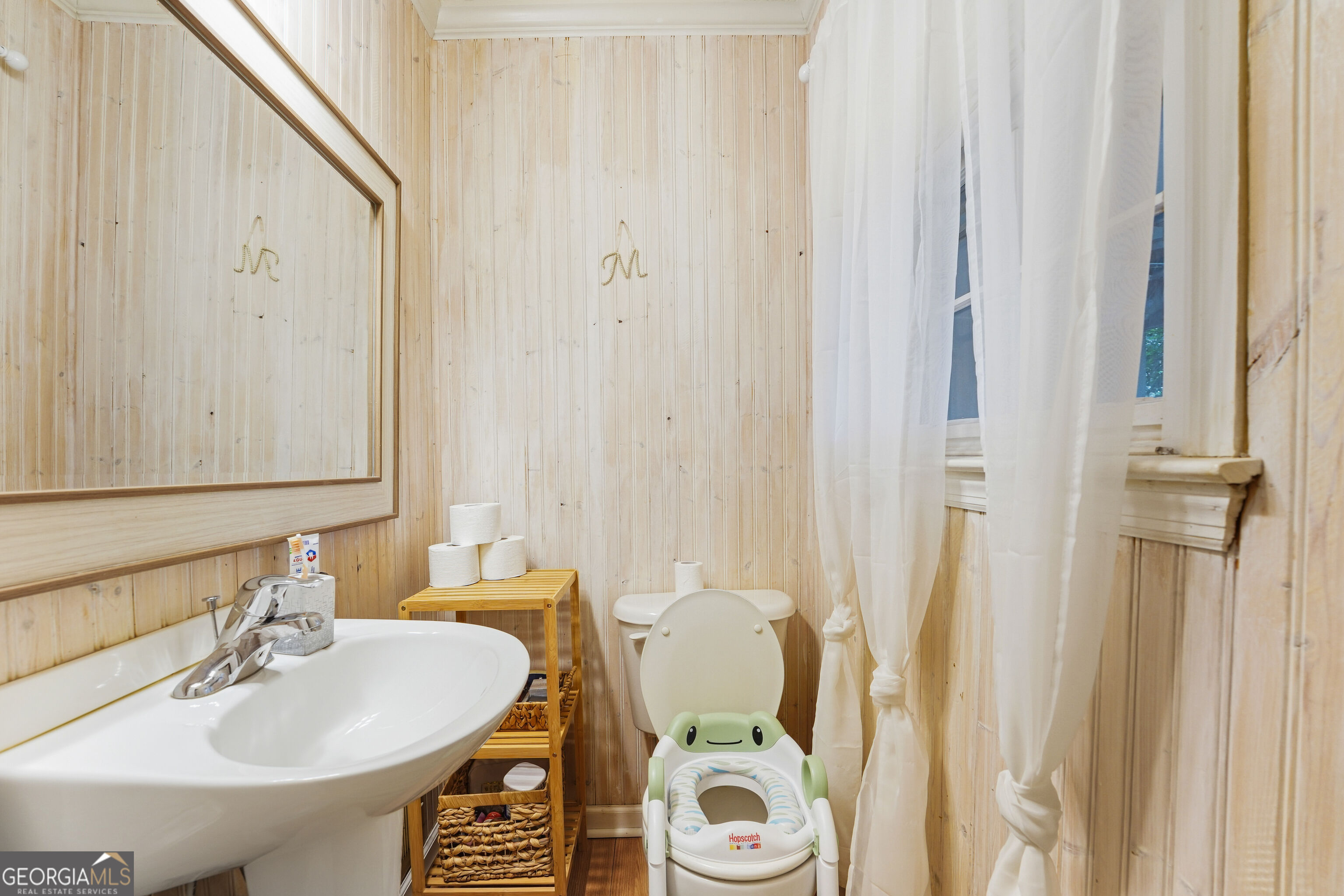 Bathroom with pale wood-paneled walls, a pedestal sink, and a toilet with a green child seat.