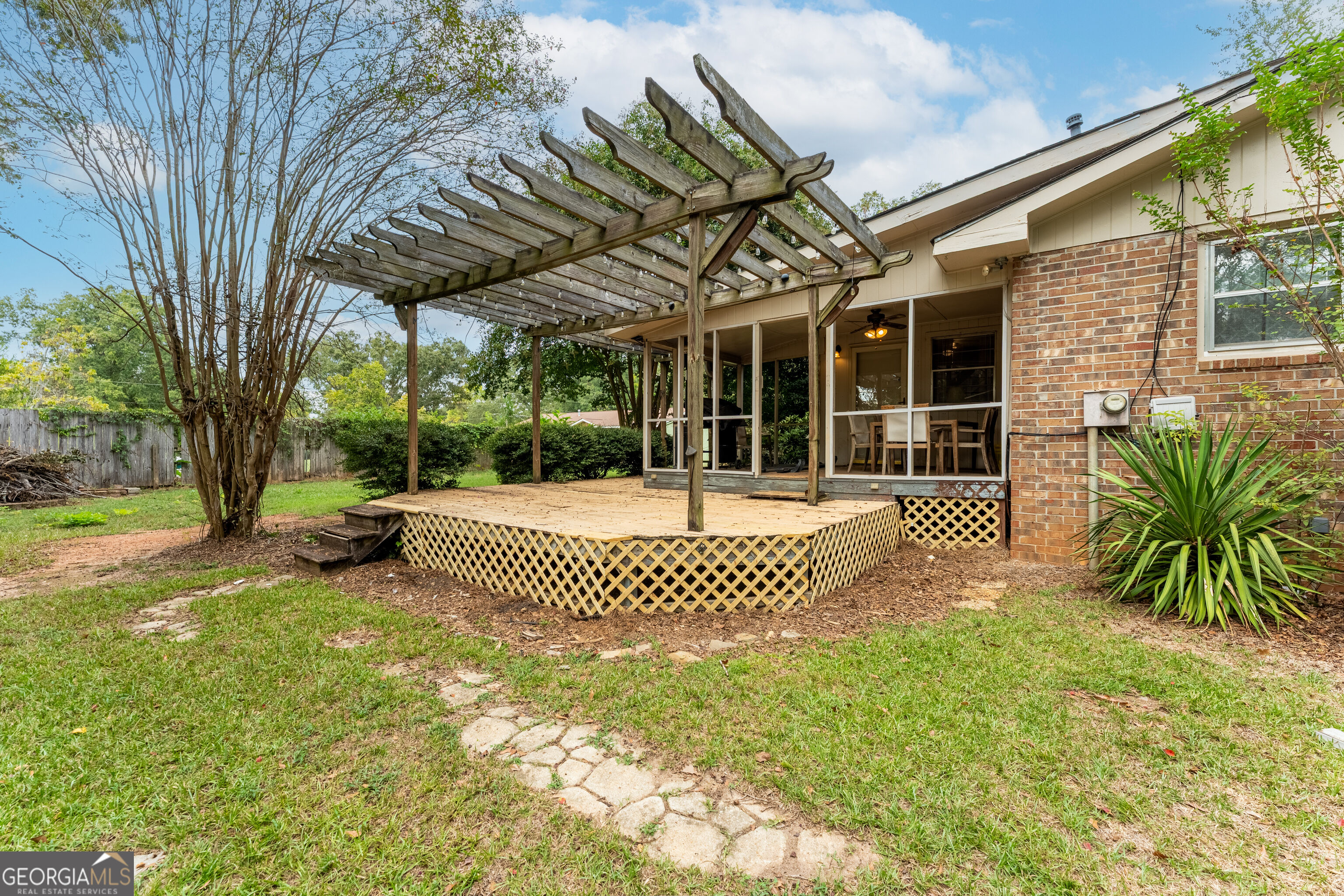 Raised wooden deck and pergola beside a brick house, with a screened porch and grassy yard.