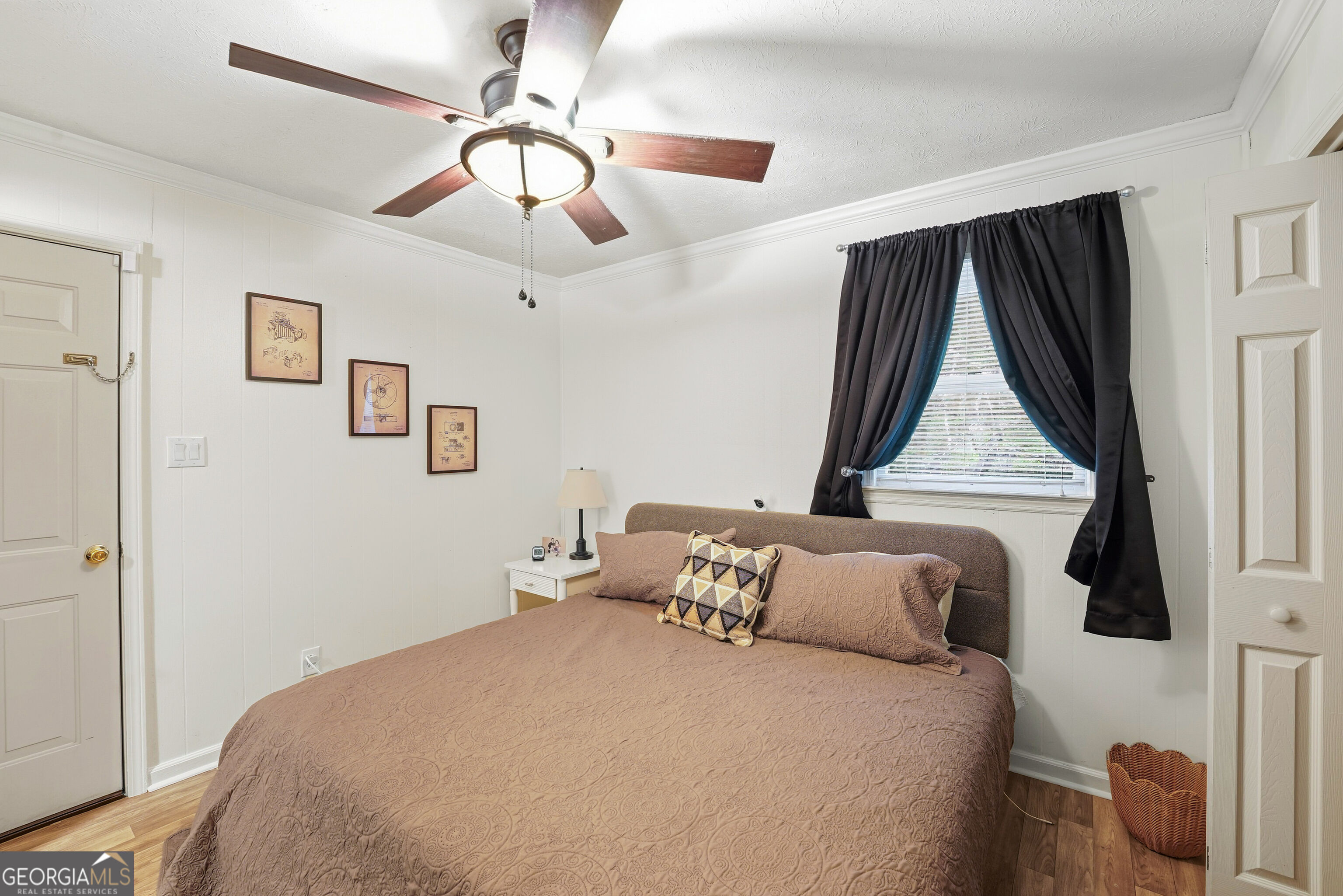 Furnished bedroom with a large bed, patterned taupe bedding, dark curtains, and a ceiling fan.