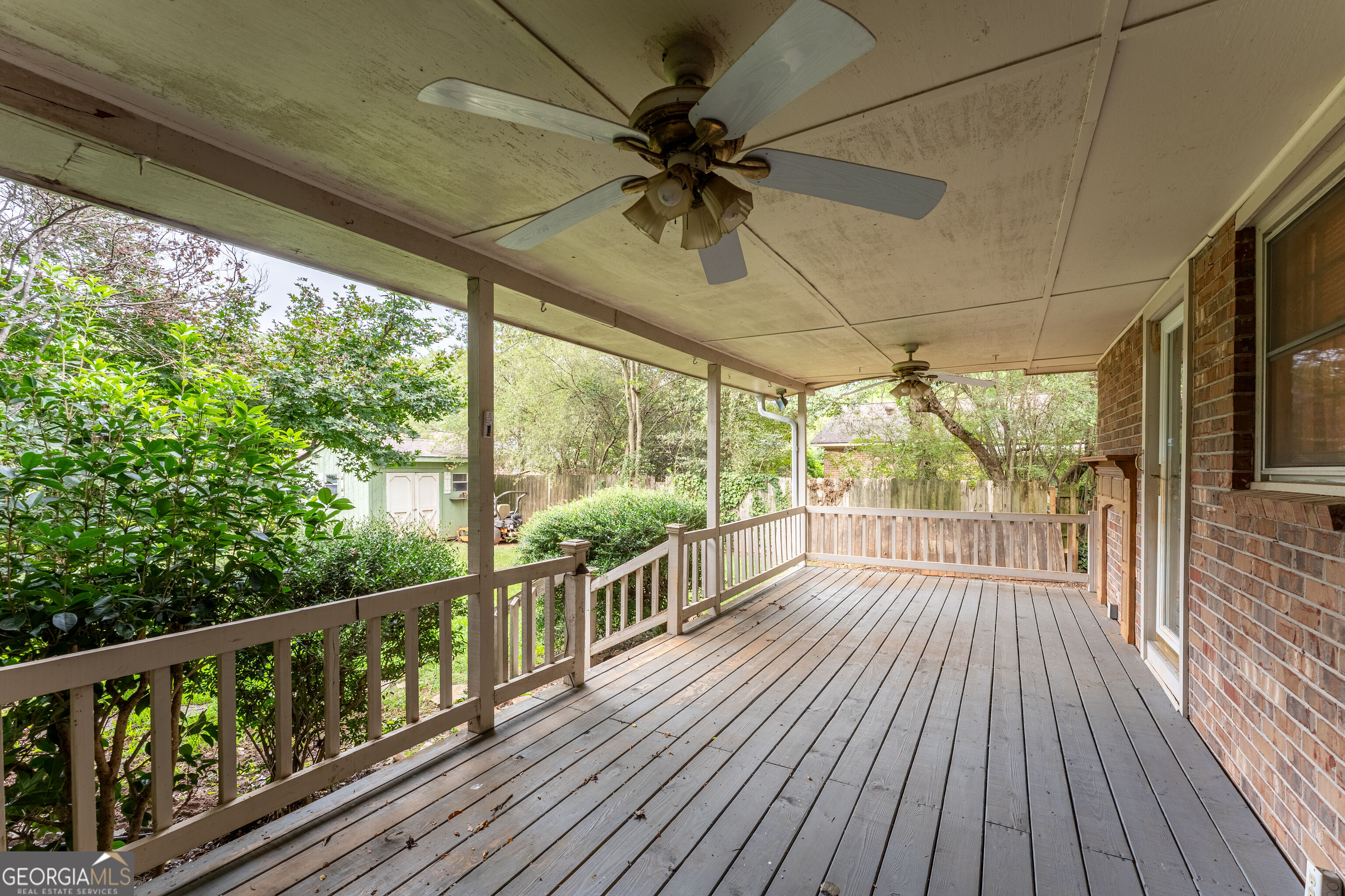 Covered wooden deck with two ceiling fans, white railings, and a leafy backyard.