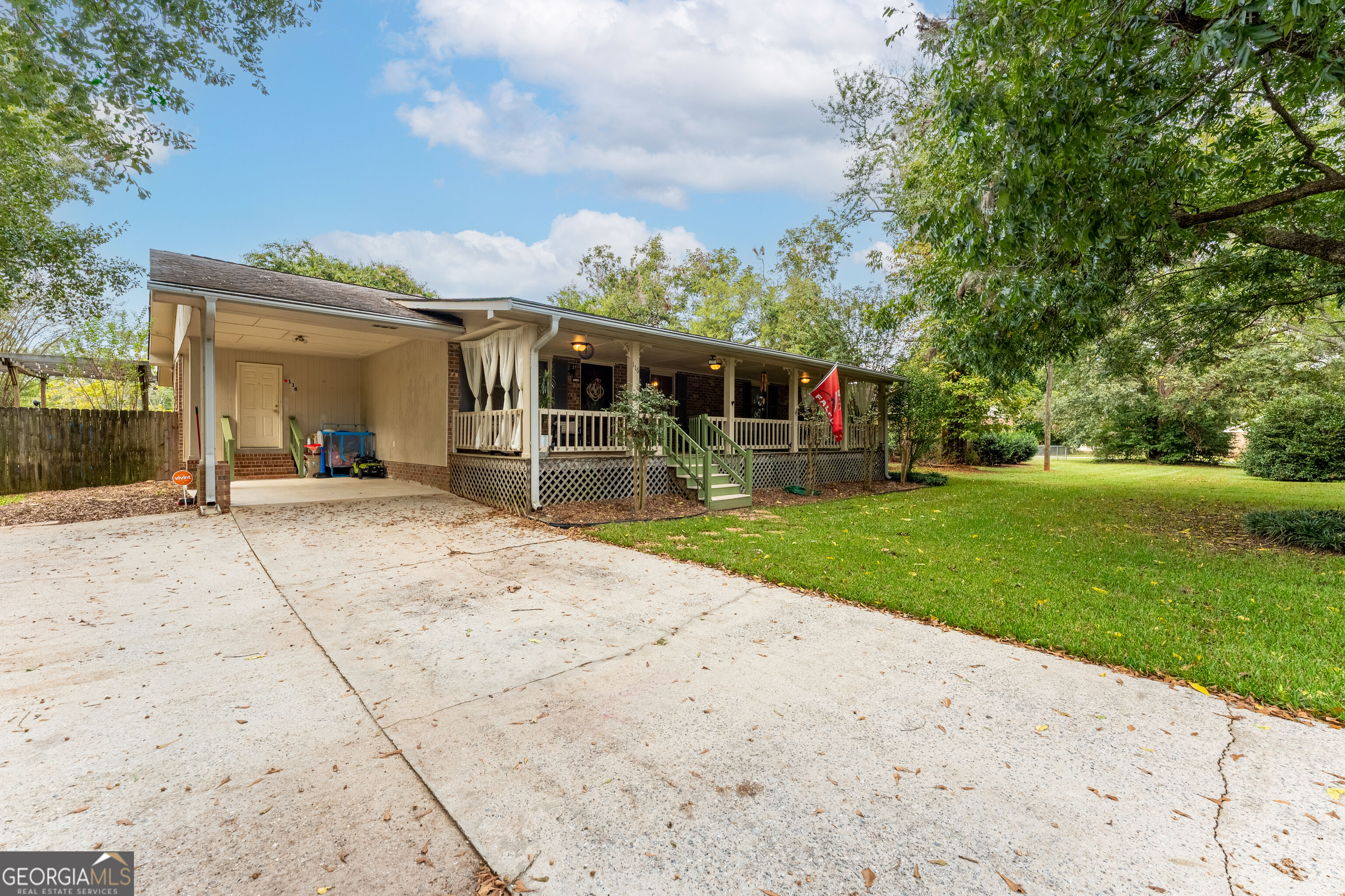 Single-story home with a long covered porch, green railings, and a wide concrete driveway beneath mature trees.