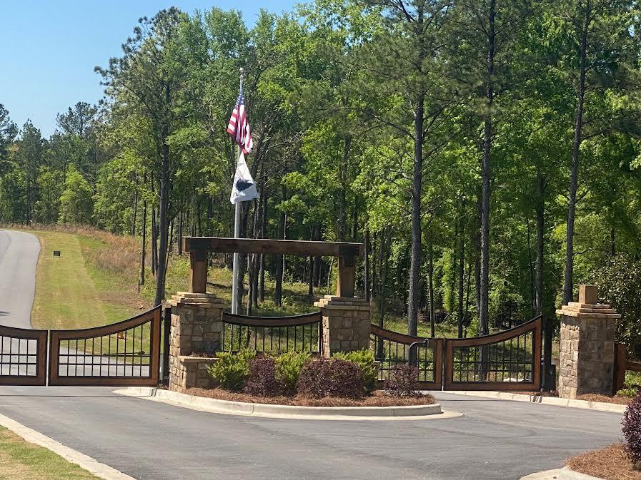Gated entrance with stone pillars, metal gates, and flags beside a wooded road.