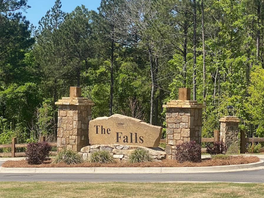 The Falls neighborhood entrance sign between stone pillars, surrounded by shrubs and pine trees.