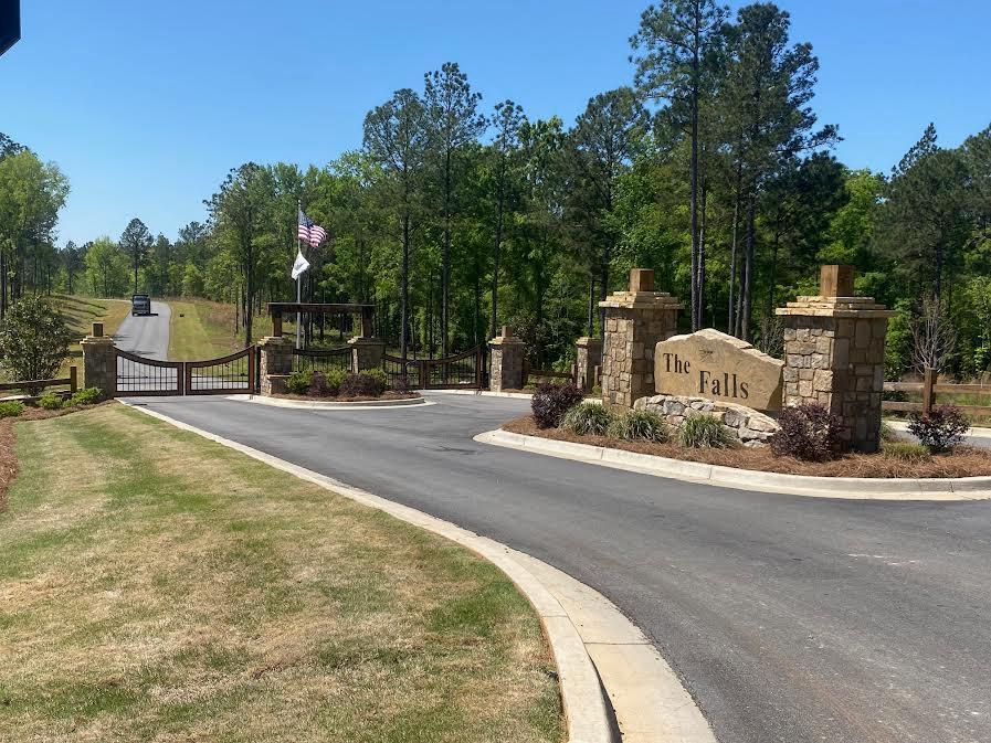 Gated entrance to The Falls, with stone pillars, a curved road, and wooded surroundings.
