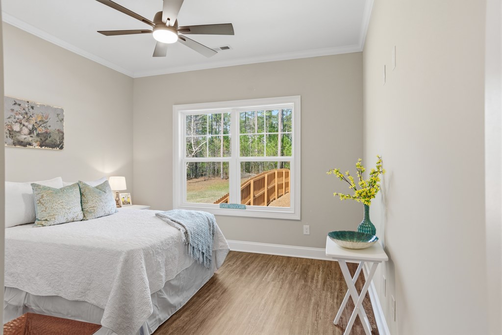Staged bedroom with a white bed, ceiling fan, and a window overlooking trees and a wooden deck.