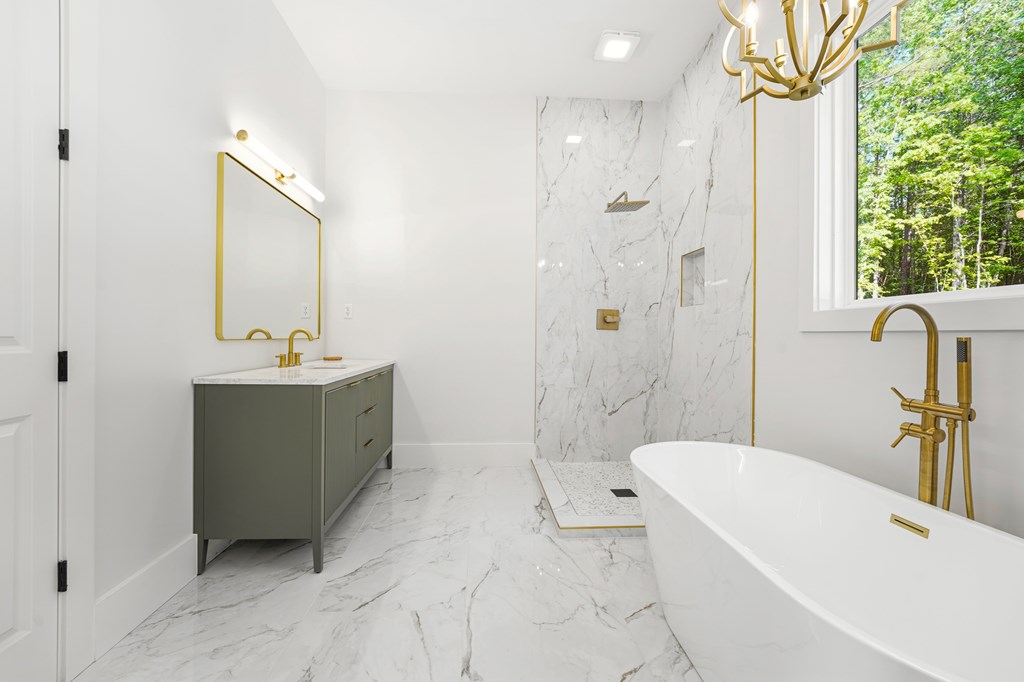 Modern bathroom with a freestanding tub, gold fixtures, marble finishes, and a window overlooking trees.