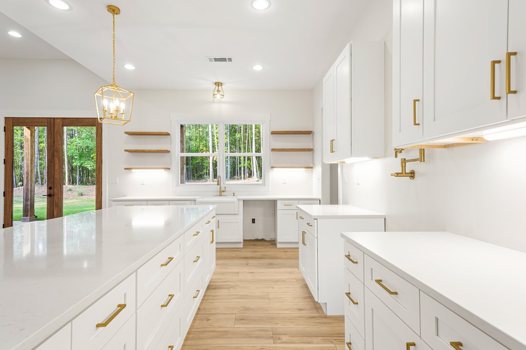 Modern white kitchen with a large island, gold fixtures, wood floors, and windows overlooking trees.
