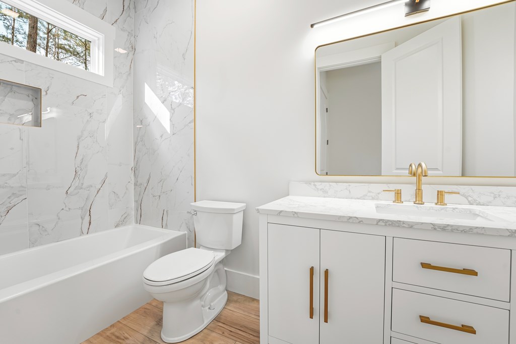 Bathroom with a marble-look tub surround, white vanity, gold fixtures, and a high window.