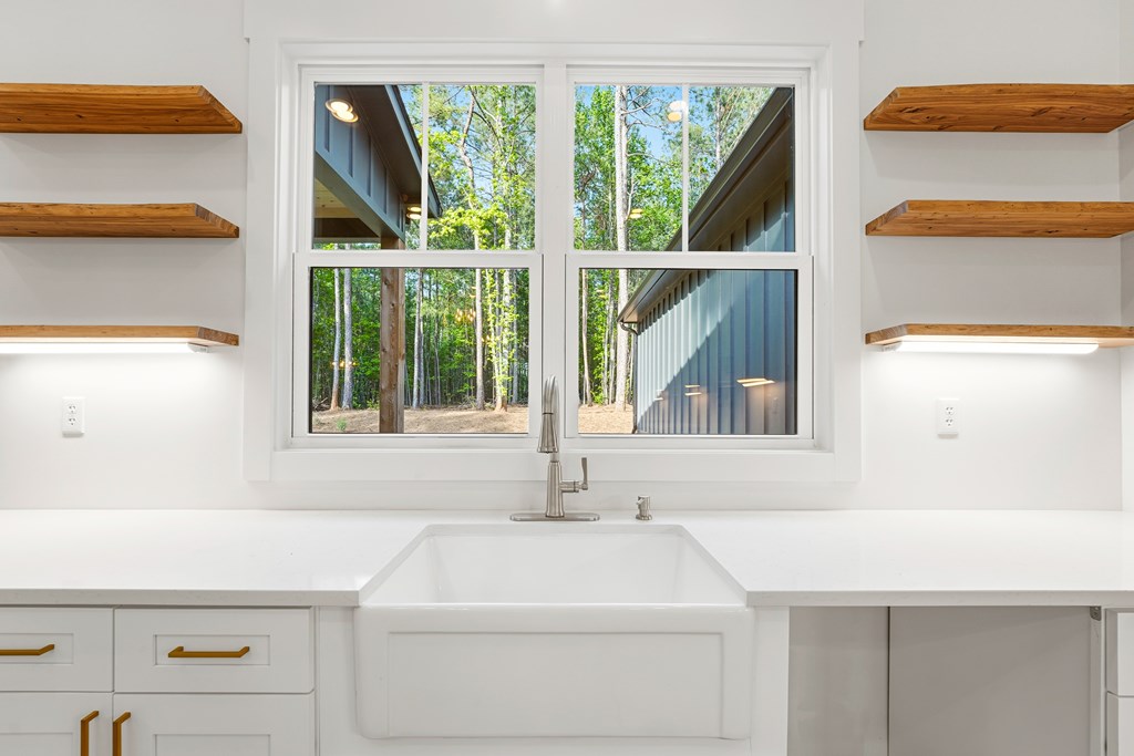 White farmhouse sink beneath a large window overlooking trees, flanked by floating wood shelves.