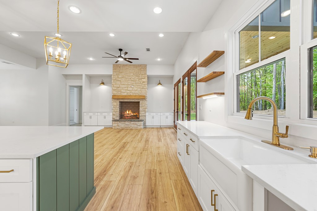 Bright kitchen with a green island, gold fixtures, and stone fireplace beside large wooded-view windows.