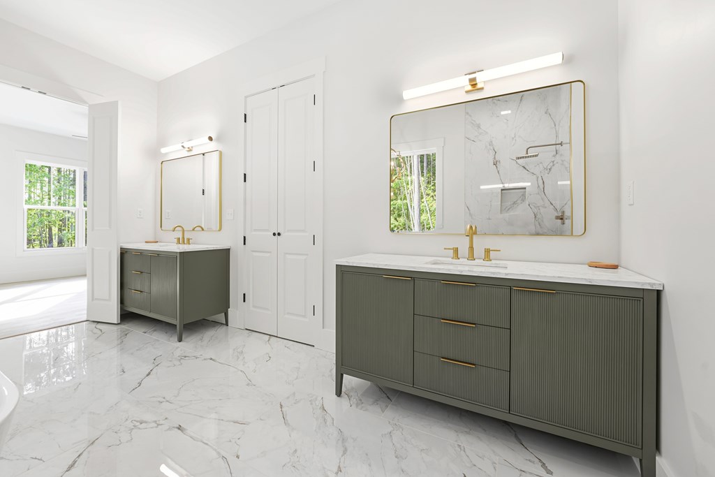 Modern bathroom with dual gray-green vanities, marble surfaces, brass fixtures, and gold-framed mirrors.