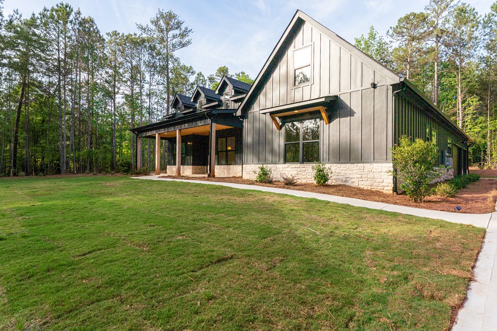 Modern farmhouse with dark siding, stone base, covered porch, and broad lawn among tall trees.