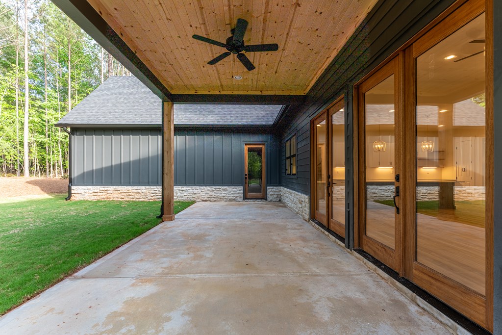 Covered concrete patio with a wood ceiling, ceiling fan, and glass doors overlooking a lawn and woods.