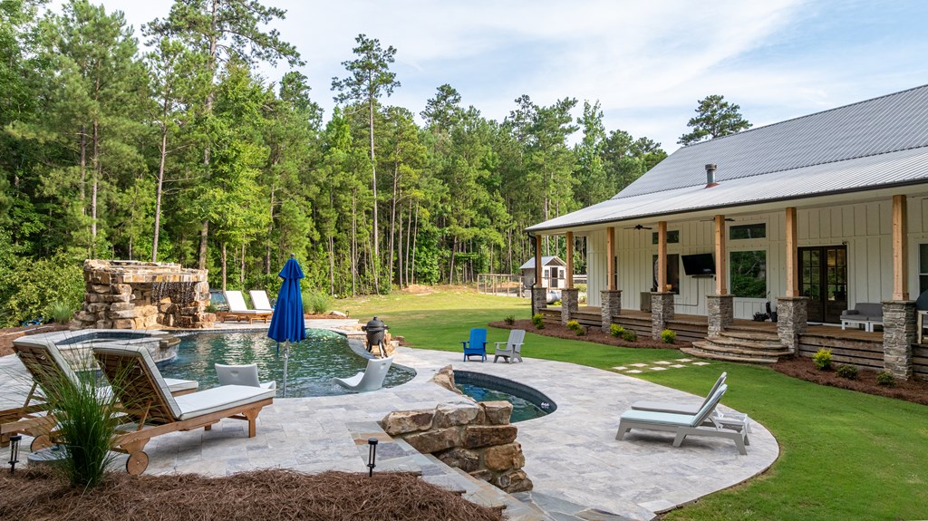 Backyard pool and stone patio beside a long covered porch, surrounded by lawn and tall trees.