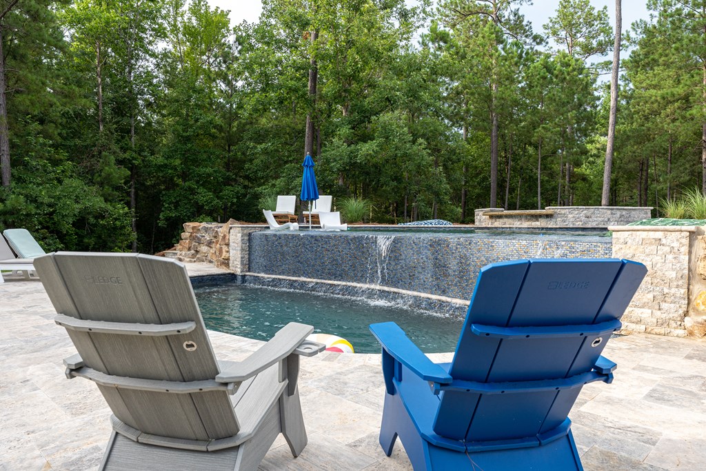 Outdoor pool with a raised waterfall wall, stone patio, lounge chairs, and wooded surroundings.