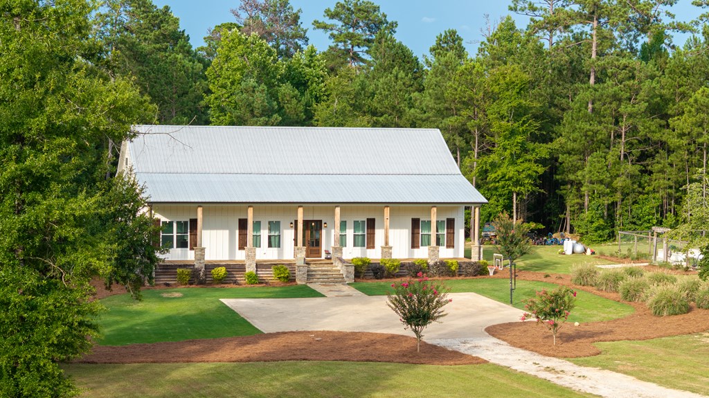 White one-story home with a tall metal roof and covered porch, set among mature trees.