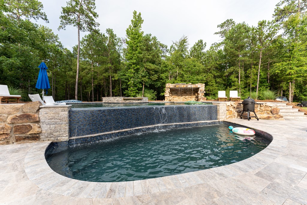 Outdoor pool with a curved basin, raised tiled edge, stone fireplace, and wooded surroundings.
