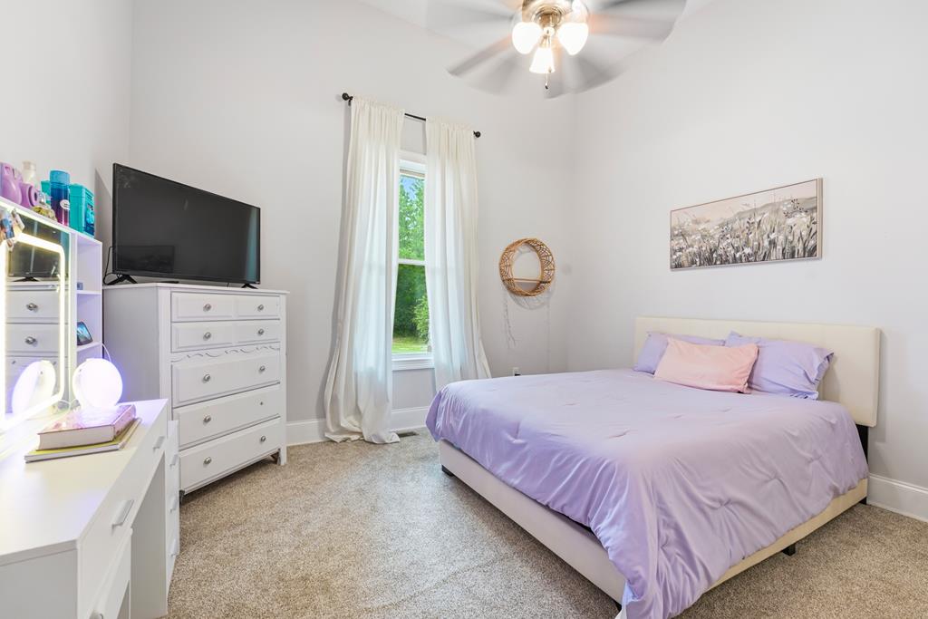 Bedroom with lavender bedding, a curtained window, ceiling fan, and television on a dresser.