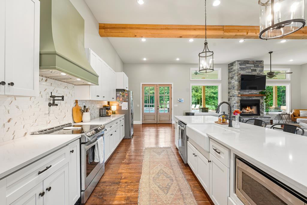 Open kitchen with a large white island, wood floors, exposed beams, and a stone fireplace beyond.