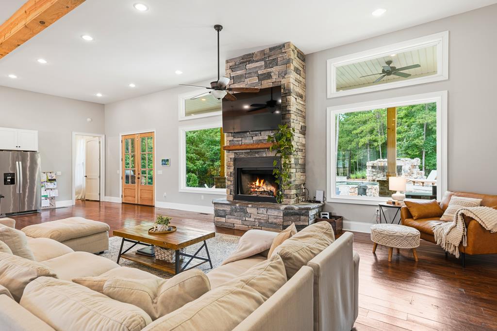 Living room with a stone fireplace, cream sectional, hardwood floors, and large windows overlooking greenery.