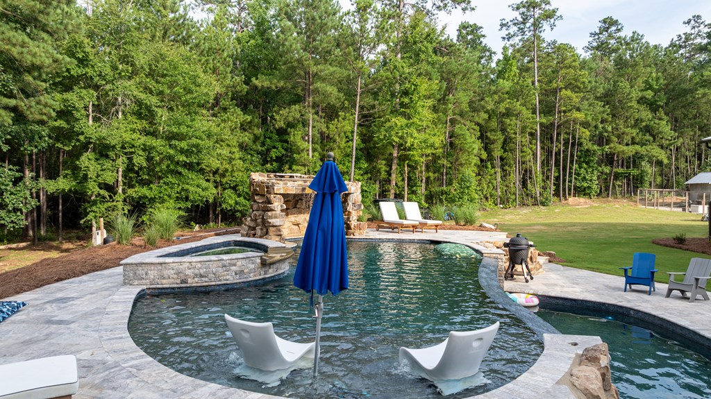 Outdoor pool with a stone waterfall, raised spa, and lounge chairs, surrounded by a wooded lawn.
