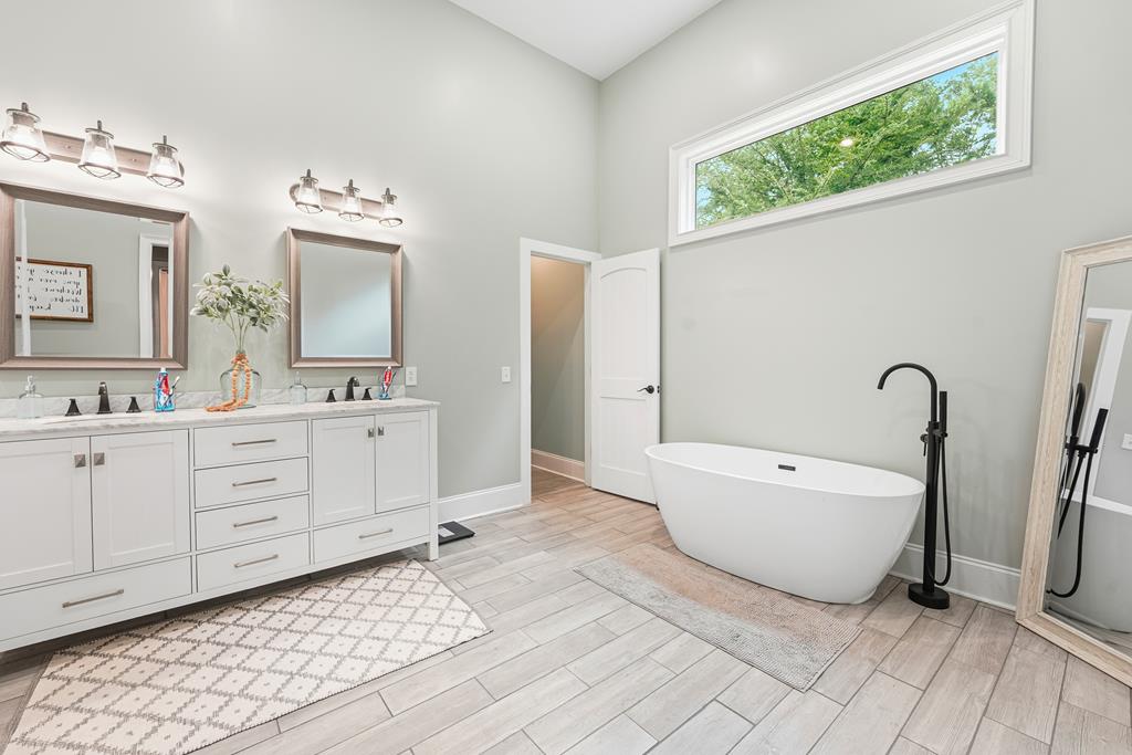 Bright bathroom with a double vanity, freestanding tub, black faucet, and high window overlooking trees.