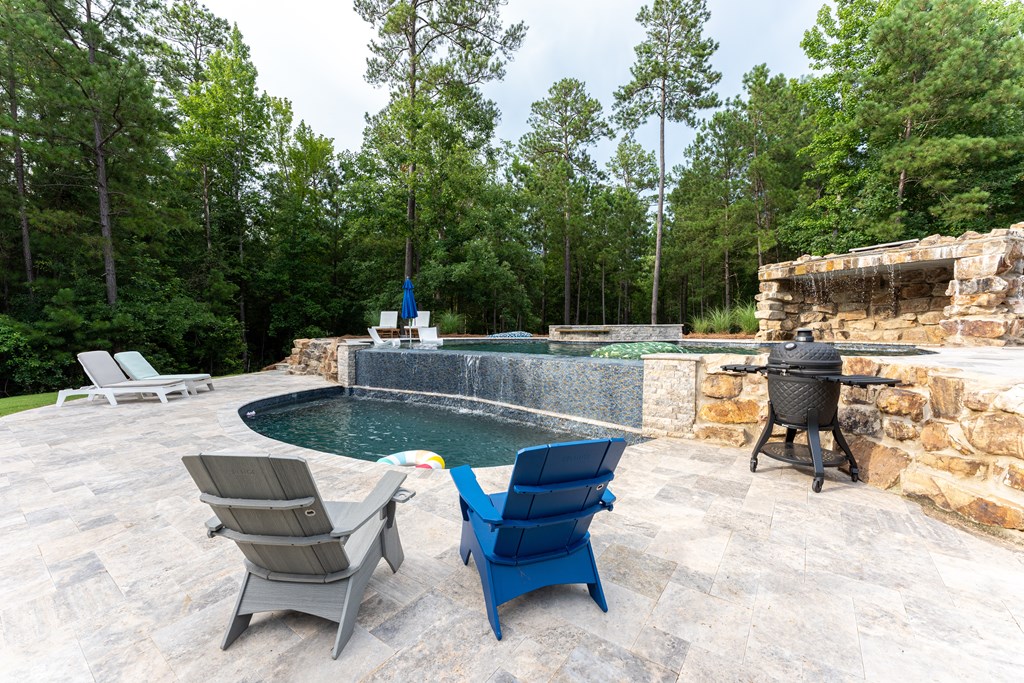 Backyard pool with a raised stone waterfall wall, tiled patio, and lounge chairs surrounded by trees.