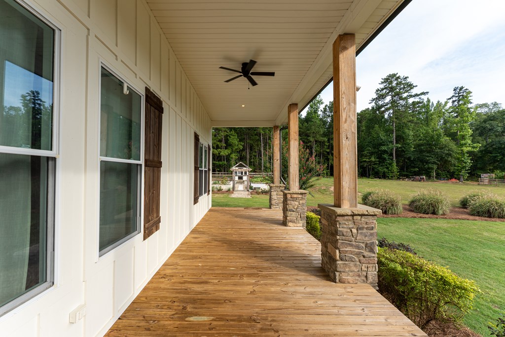 Covered wooden porch with a ceiling fan and stone-based posts overlooking a landscaped lawn and trees.