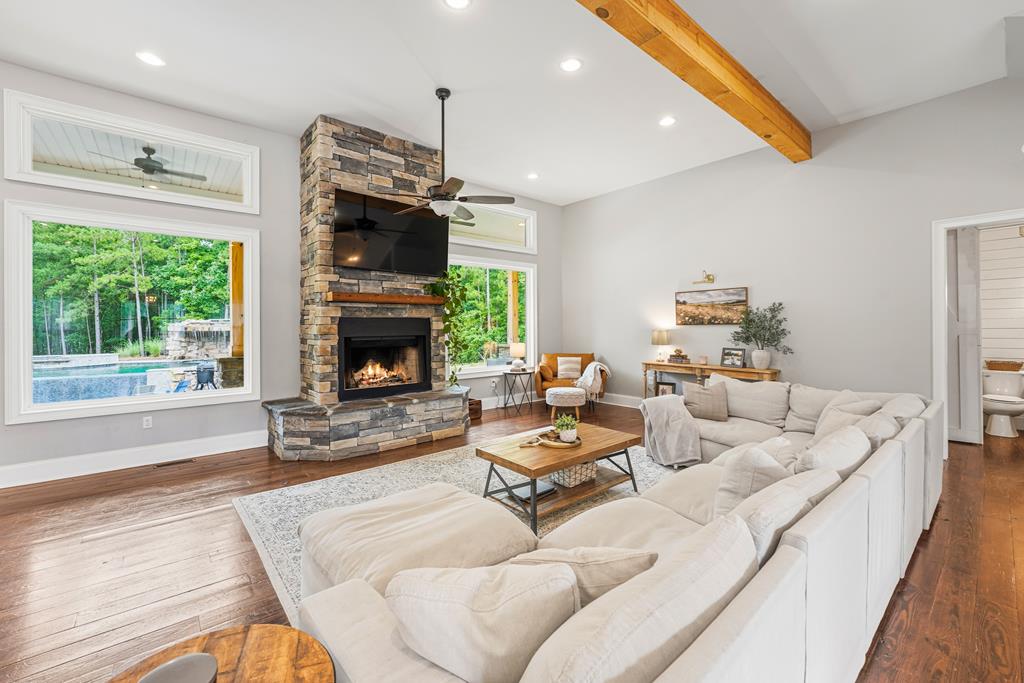 Living room with a stone fireplace, white sectional, exposed ceiling beam, and windows overlooking a pool.
