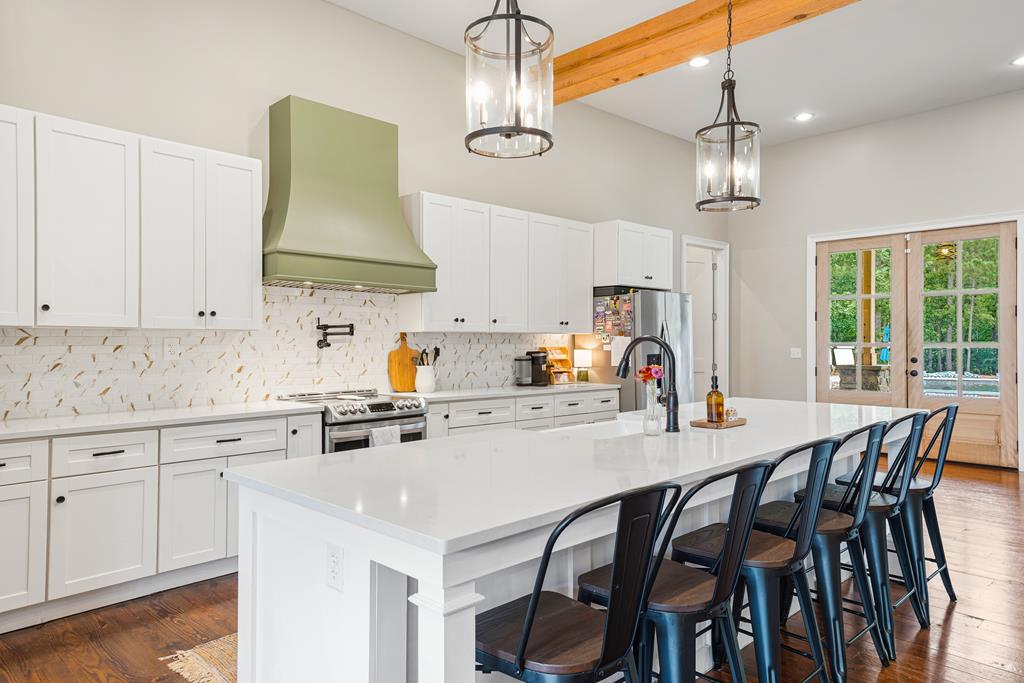 Kitchen with a large white island, dark stools, green range hood, and glass doors to a wooded view.