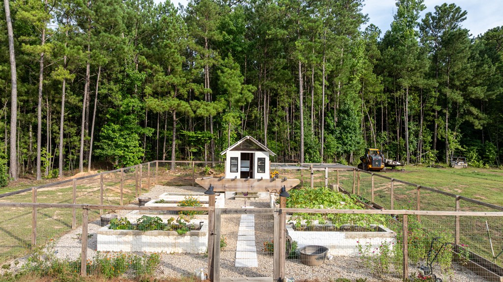 Fenced vegetable garden with raised beds and gravel paths, backed by woods and a small white shed.