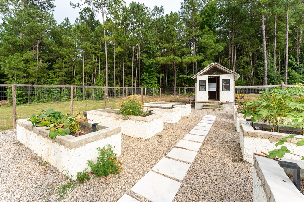 Fenced garden with white raised beds, a gravel path, and a small shed among pine trees.