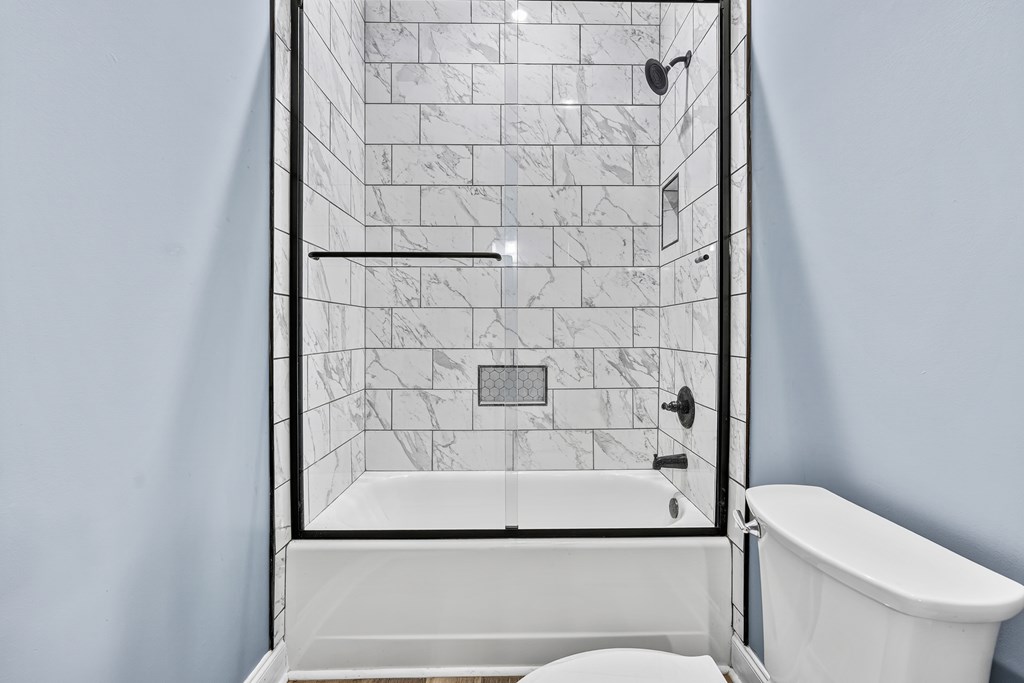 Bathroom with a marble-look tiled shower, glass enclosure, white tub, and dark fixtures.