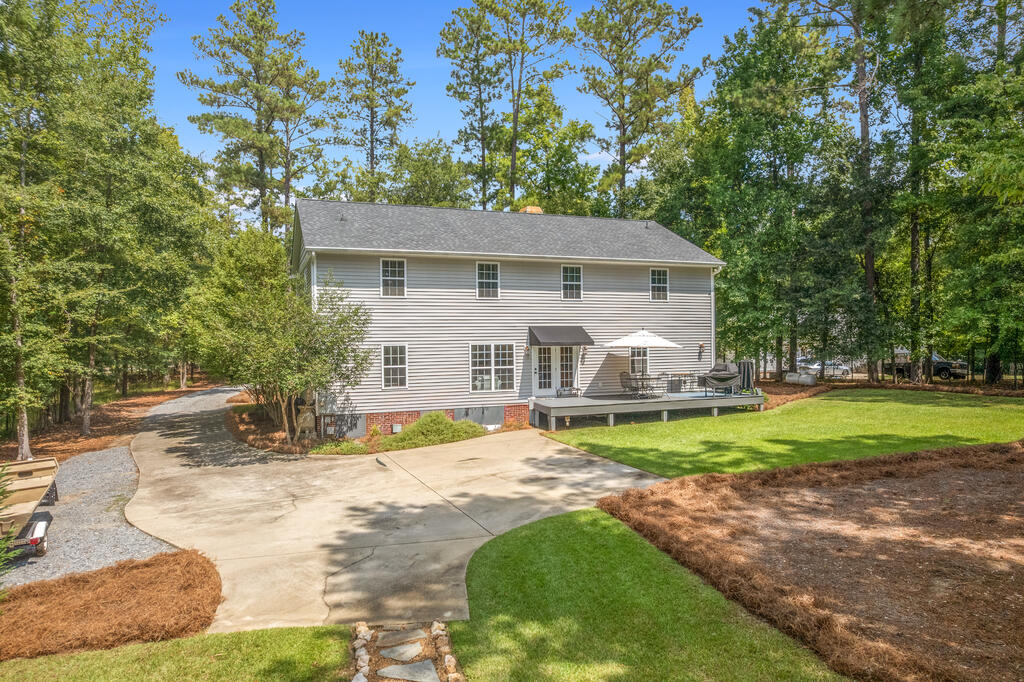 Two-story gray house with a raised outdoor deck, broad concrete patio, and wooded surroundings.