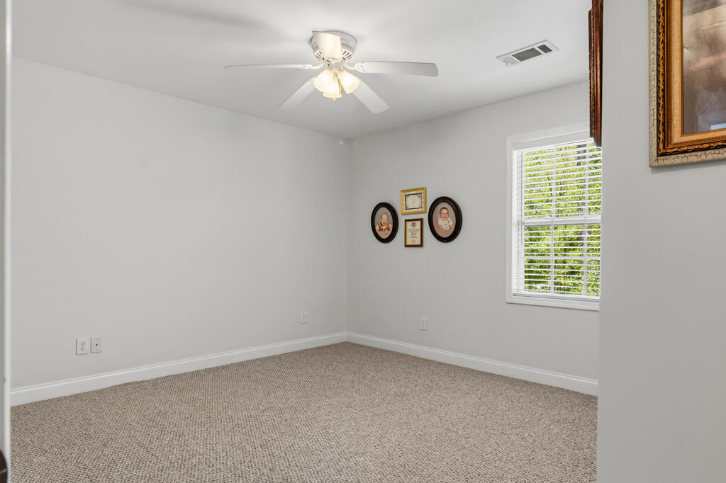 Vacant carpeted bedroom with a ceiling fan, white blinds, and framed pictures.