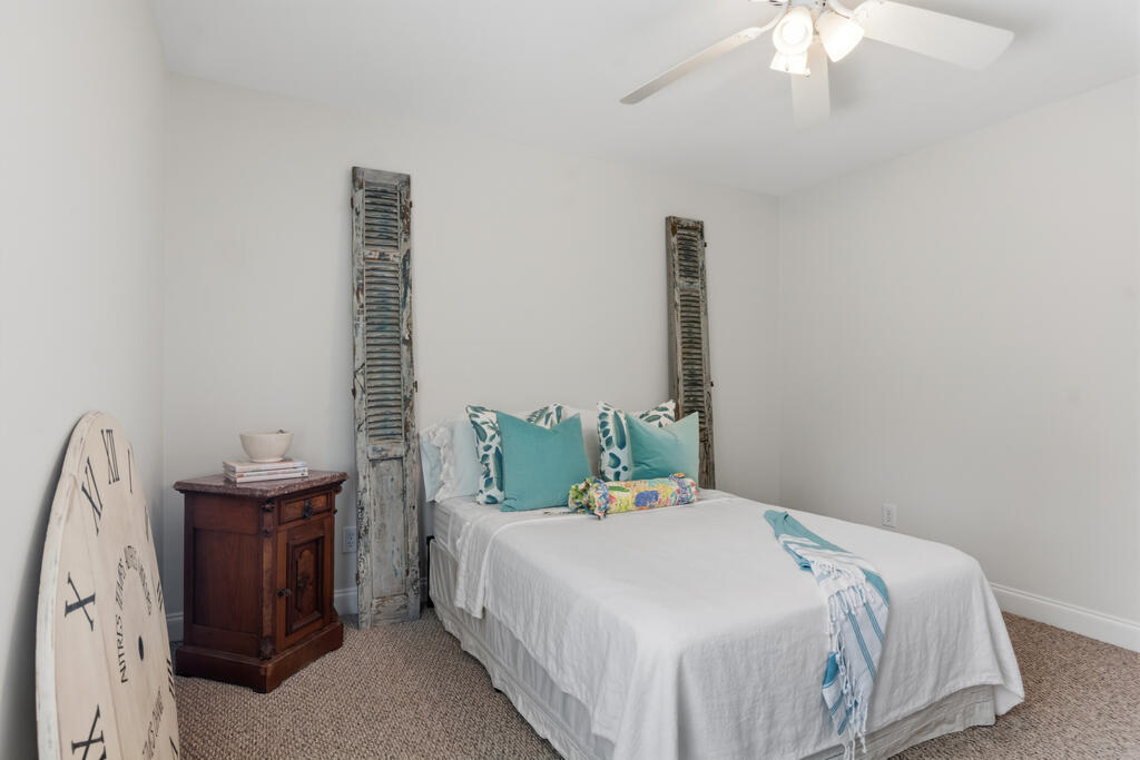 Staged bedroom with a white bed, turquoise pillows, weathered shutters, and a ceiling fan.