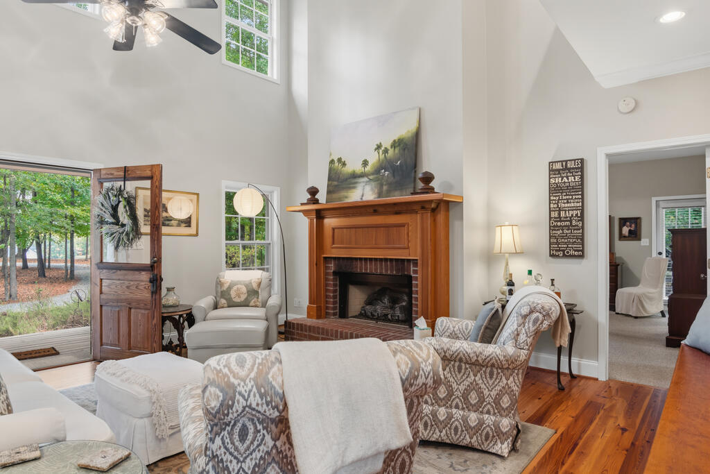 Living room with a tall vaulted ceiling, wooden fireplace mantel, patterned armchairs, and an open front door.