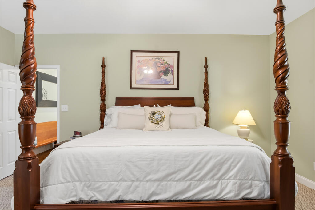 Bedroom with a large four-poster bed, white bedding, floral artwork, and a lit bedside lamp.