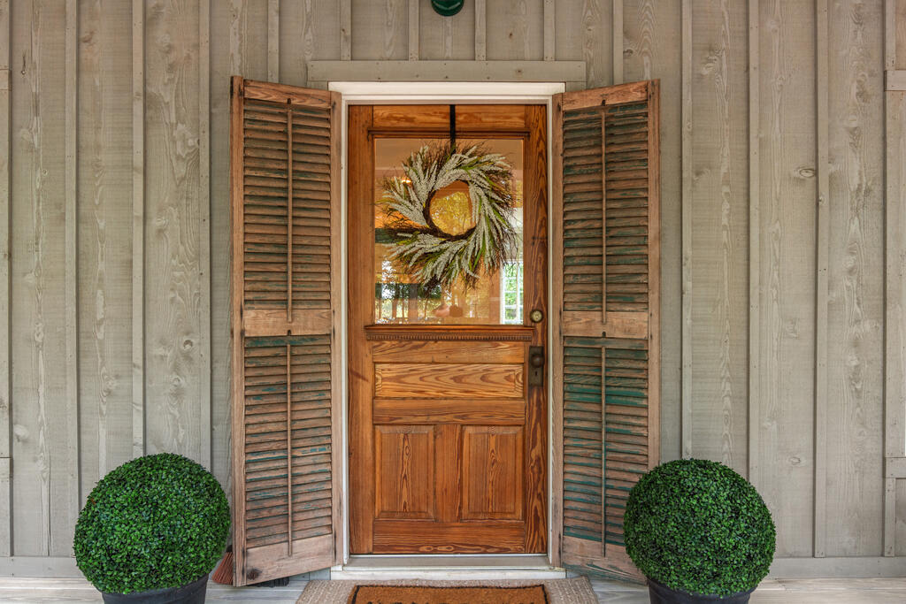 Wooden front door with a wreath, open shutters, and potted topiary shrubs.