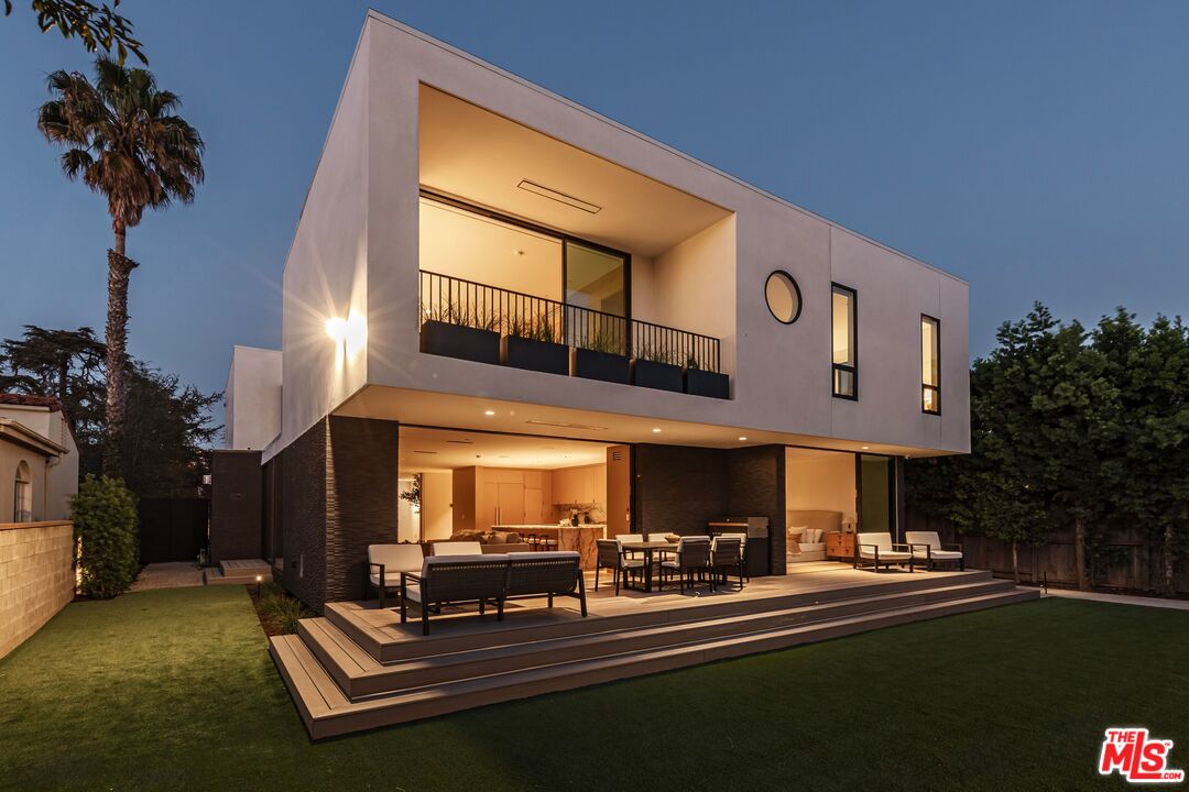 Modern two-story home with a white facade, illuminated patio, tiered deck, and lawn at dusk.