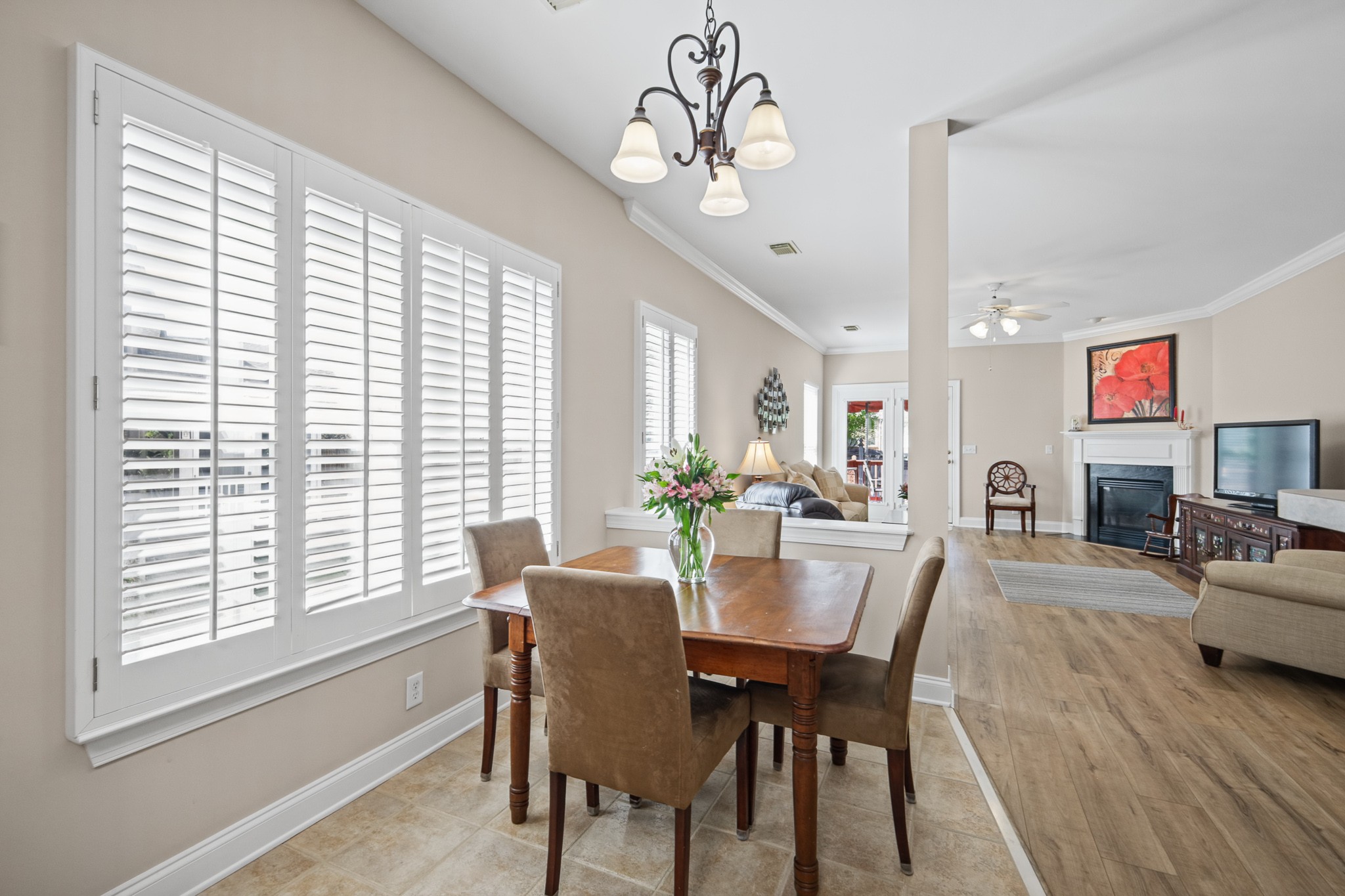 Dining room with a wooden table and plantation shutters opening into a living room with a fireplace.