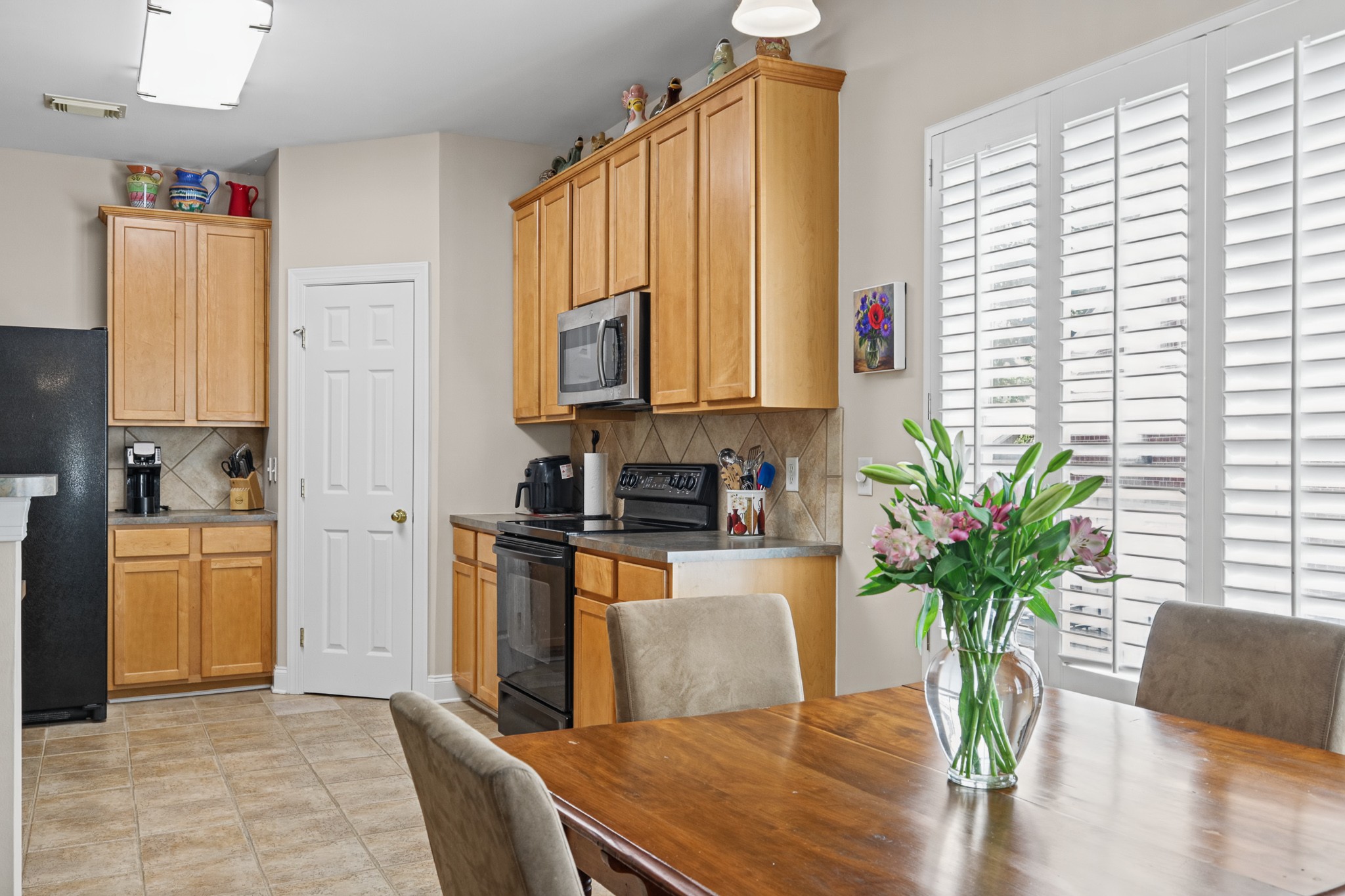 Kitchen with honey-colored cabinets, black appliances, and a dining table beside shuttered windows.