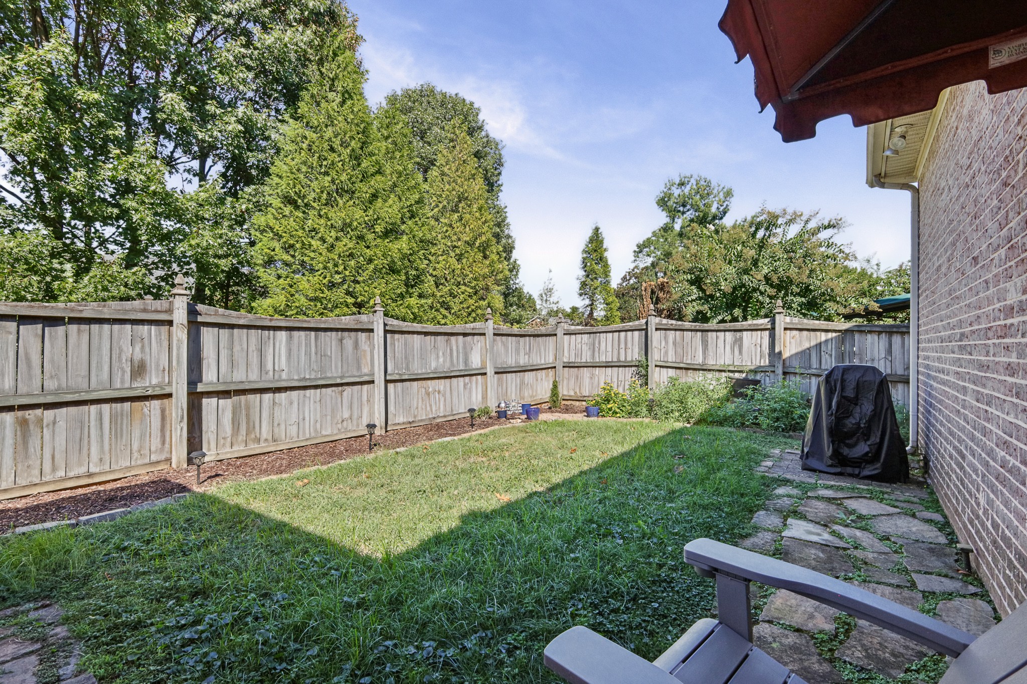 Fenced backyard with a grassy lawn, stone path, and covered grill beside a brick house.