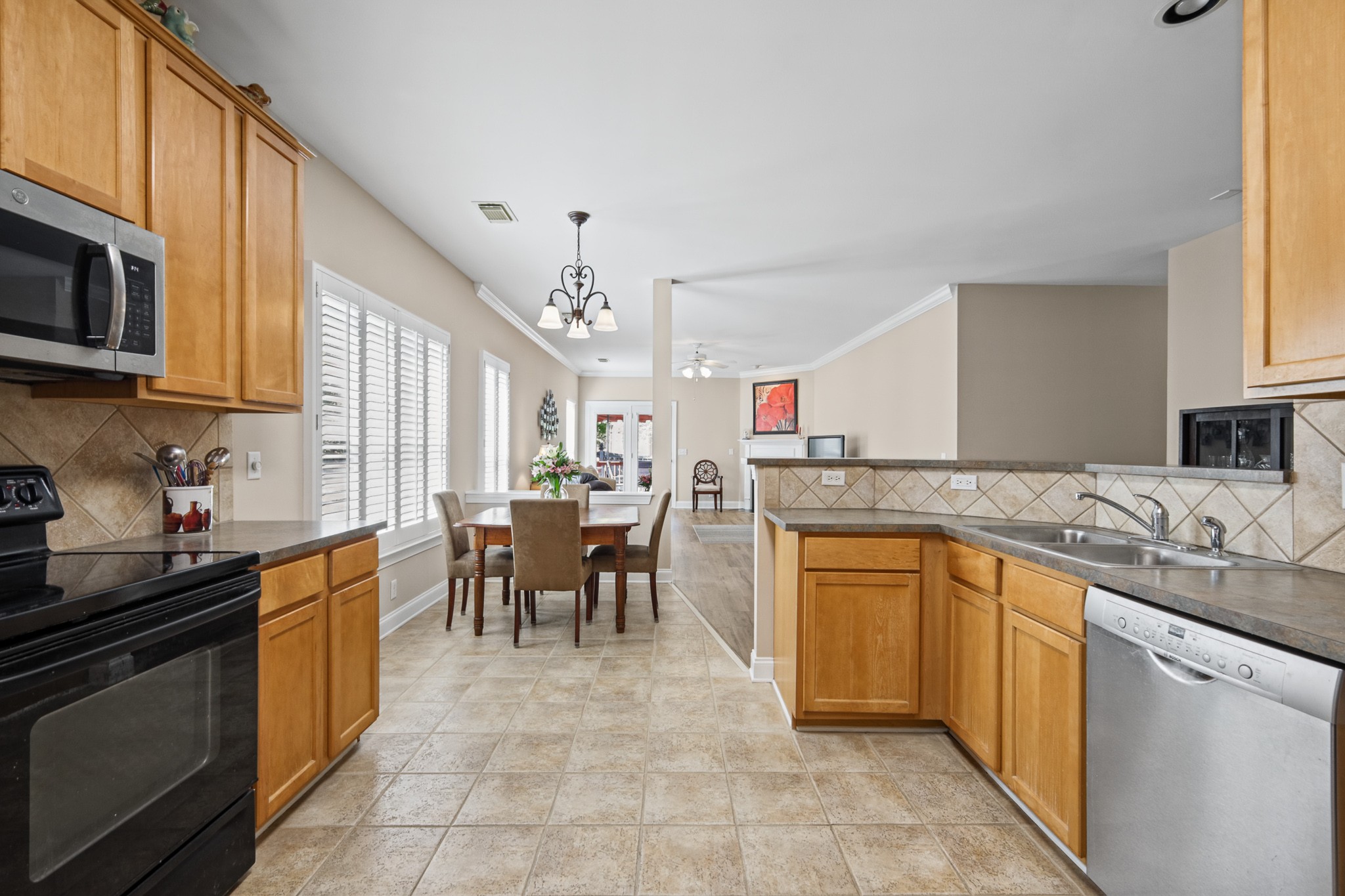 Kitchen with honey-colored cabinets and tile floors opening to a dining area by shuttered windows.