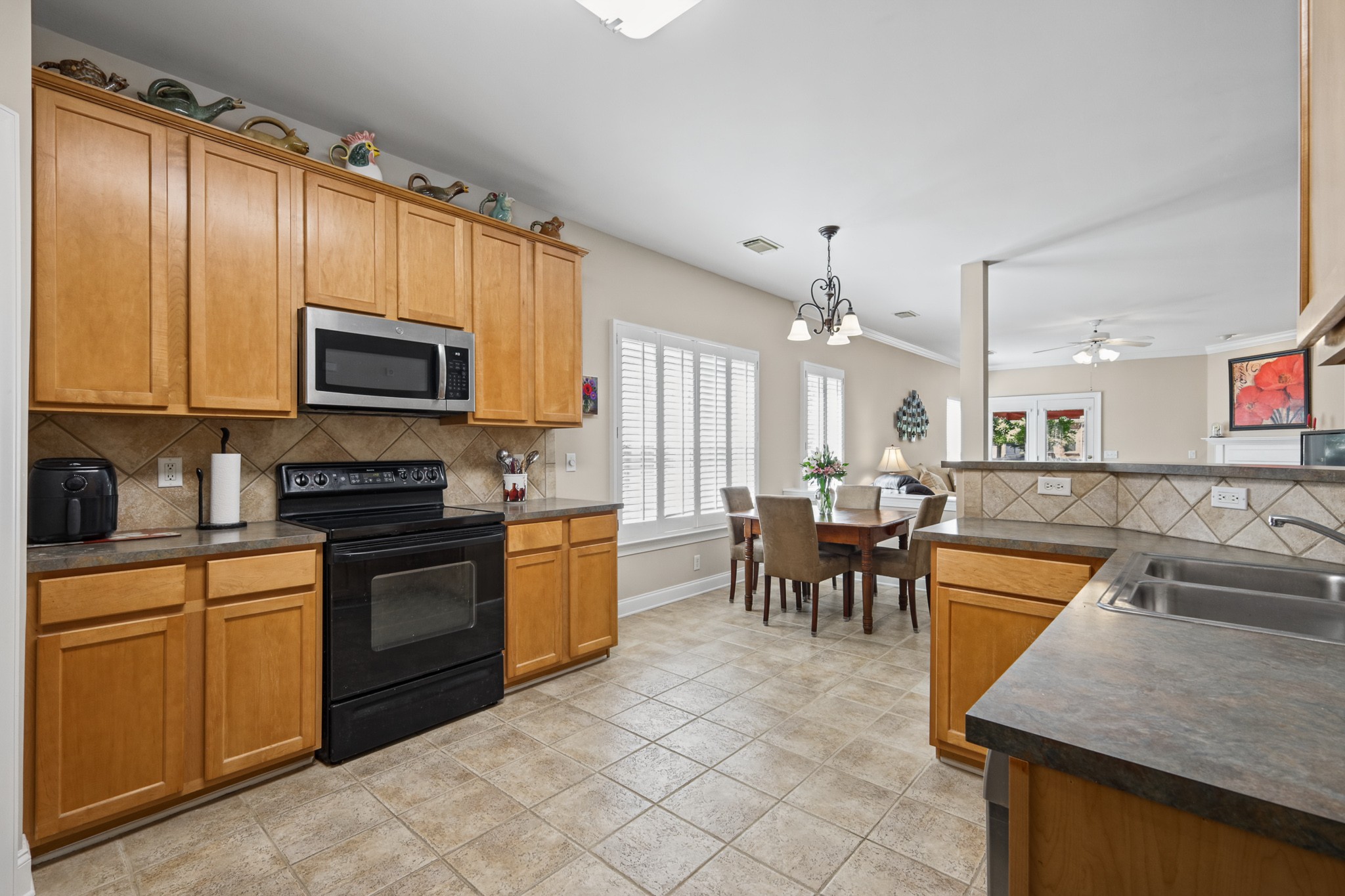 Kitchen with honey-colored cabinets and black appliances, opening to a dining area by shuttered windows.