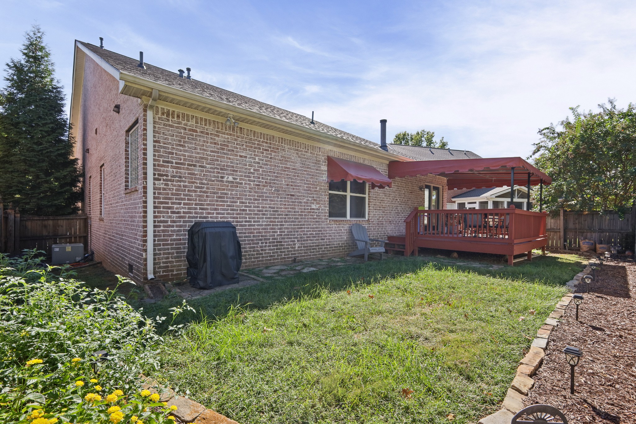 Brick house backyard with a red deck and awning, fenced lawn, and garden beds.
