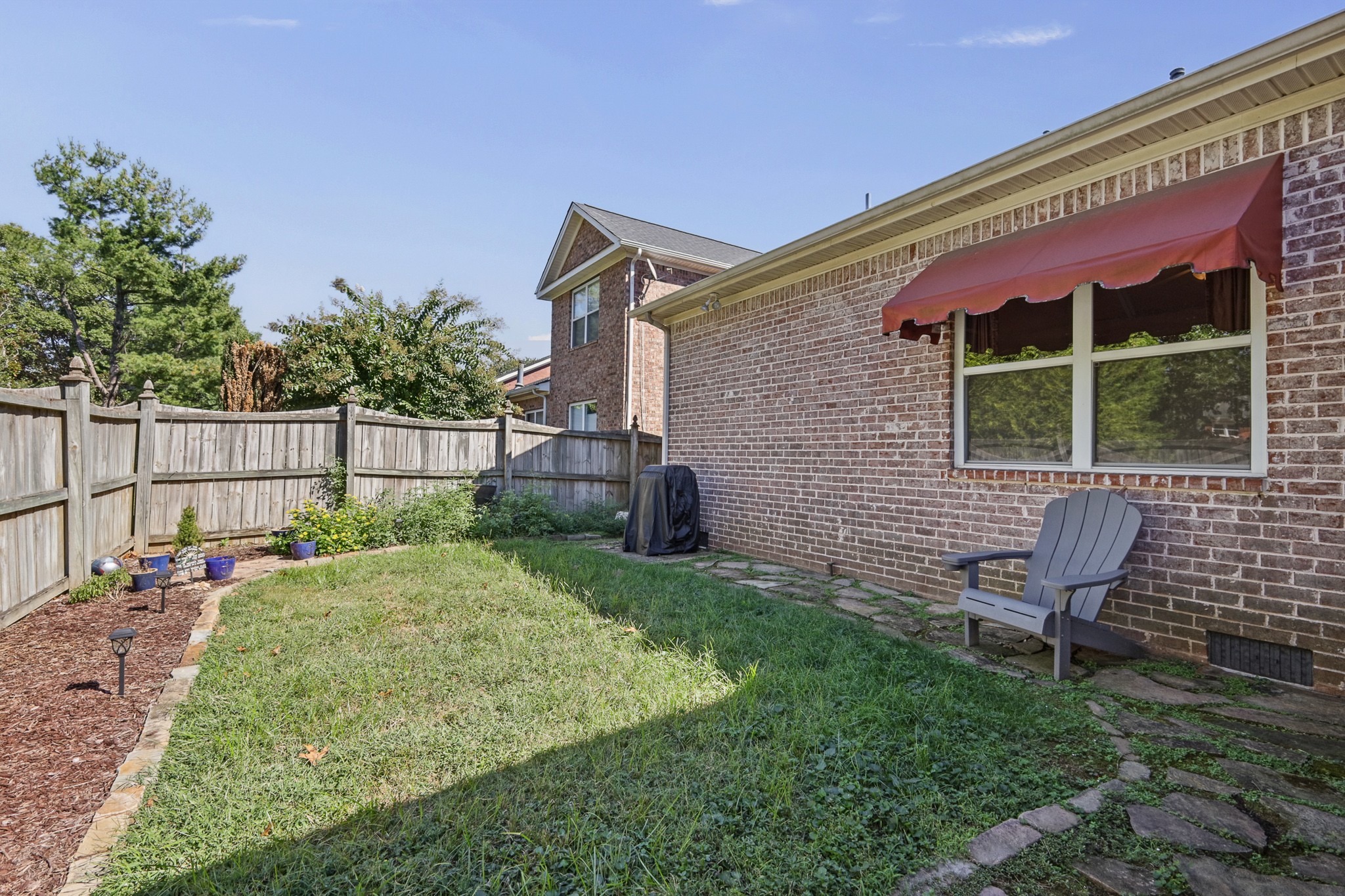 Fenced backyard with a grassy lawn, stone path, garden beds, and a chair beside the brick house.