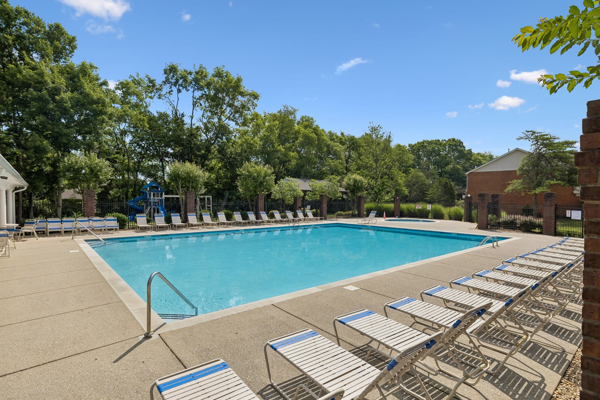 Outdoor swimming pool with rows of lounge chairs, fenced deck, and a playground beyond.