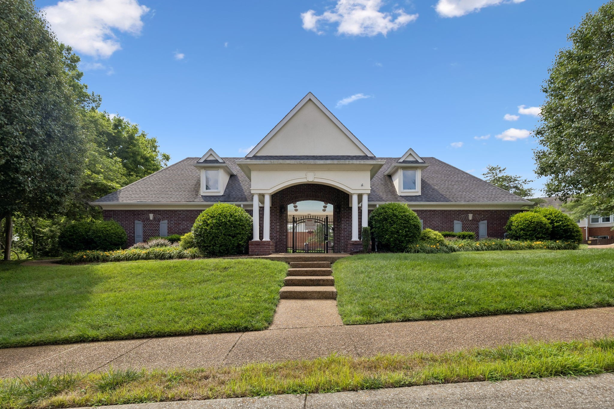 Brick community entrance with an arched black gate, white columns, and a manicured lawn.